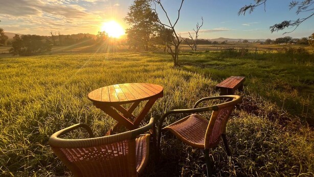 A wooden table and two wicker chairs in a grassy field with a sunset in the background. - Vacy Hunter Valley Lodge (Vacy)