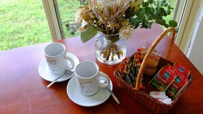 Una mesa con dos tazas blancas y platillos, una cuchara, un florero de vidrio con flores secas y una cesta de bocadillos.