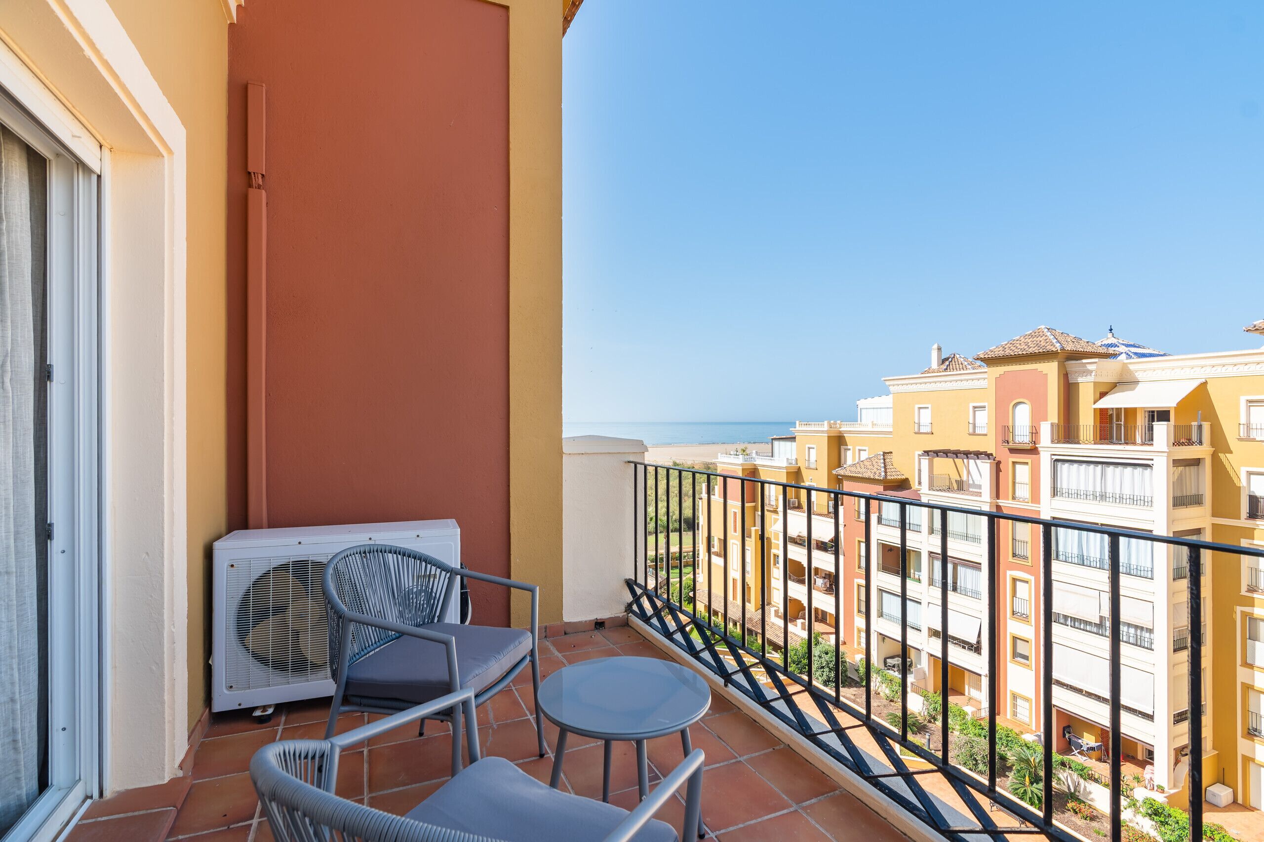 Un balcón con mesa y sillas, un equipo de aire acondicionado y vistas a edificios cercanos.