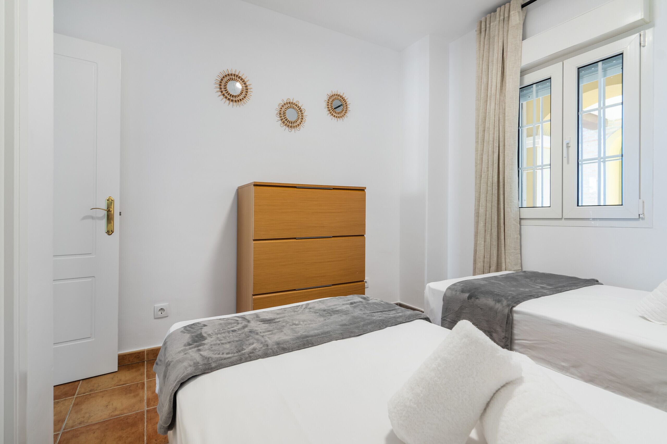 Ein Zimmer mit zwei Betten, einem Holzkasten, einer weißen Tür und einem Fenster mit Vorhängen.