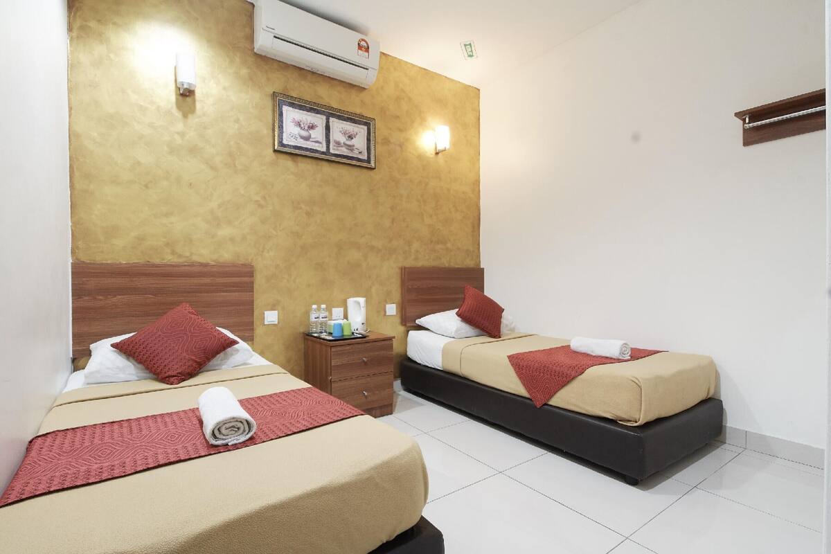 A hotel room with two beds, each with a red pillow, a beige bedspread, and a rolled towel. There is a wooden nightstand with a lamp and a framed picture on the wall.