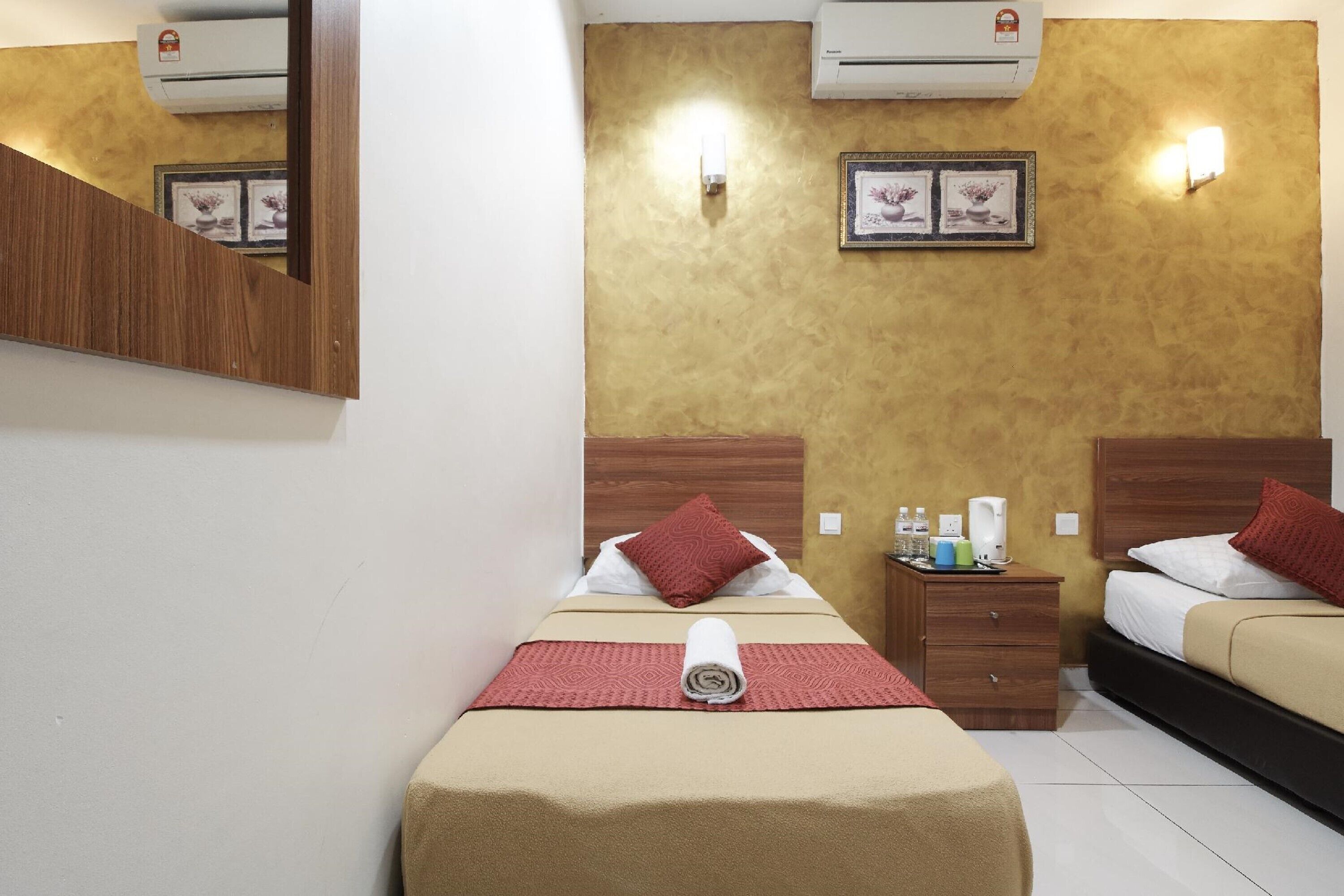 A hotel room with two beds, a wooden headboard, a small bedside table with toiletries, and a wall-mounted air conditioner.