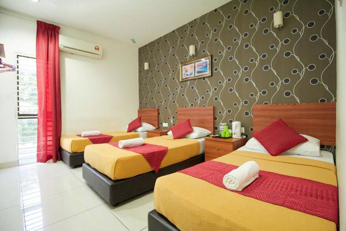 A hotel room with three beds, each with a red pillow and a yellow bedspread. There is a window with red curtains, an air conditioning unit on the wall, and a framed picture above the beds.