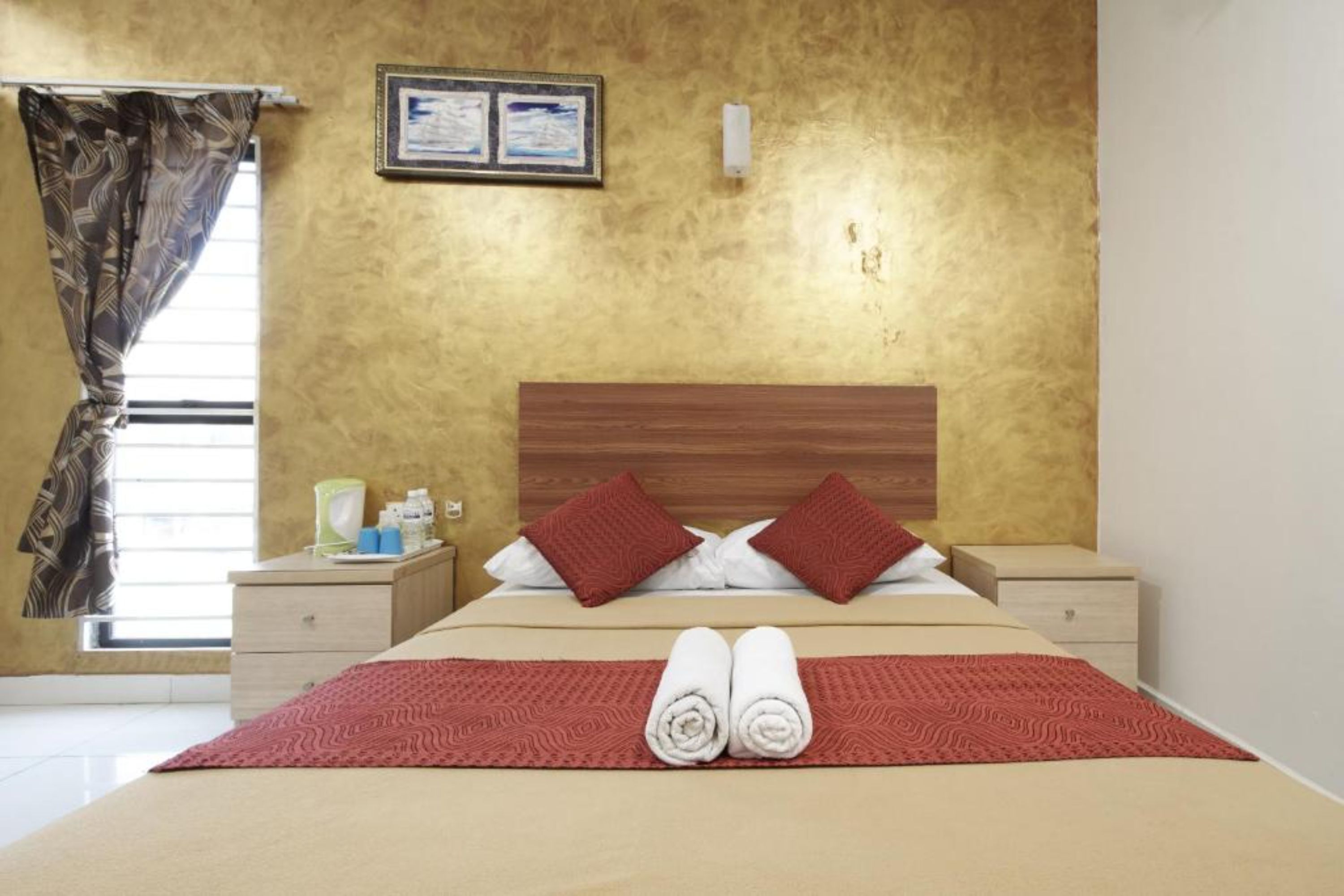 A hotel room with a bed, two pillows, a red blanket, a wooden headboard, a nightstand, a lamp, and a window with curtains.