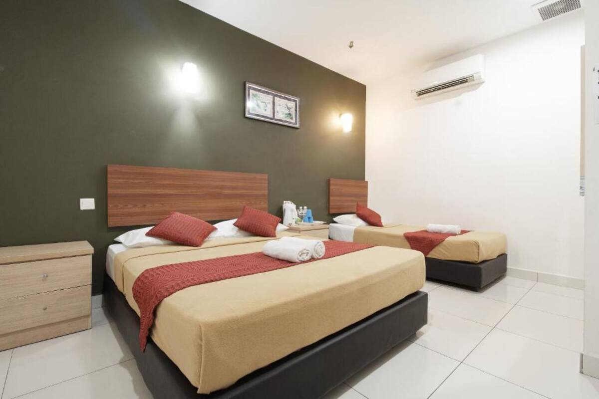 A hotel room with two beds, each with a wooden headboard, beige bedding, and red pillows. There is a nightstand with toiletries, a framed picture on the wall, and an air conditioning unit.