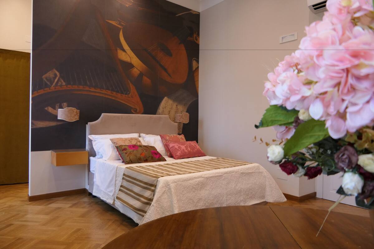 A bedroom with a bed, pillows, a nightstand, a wall mural, and a vase of flowers.