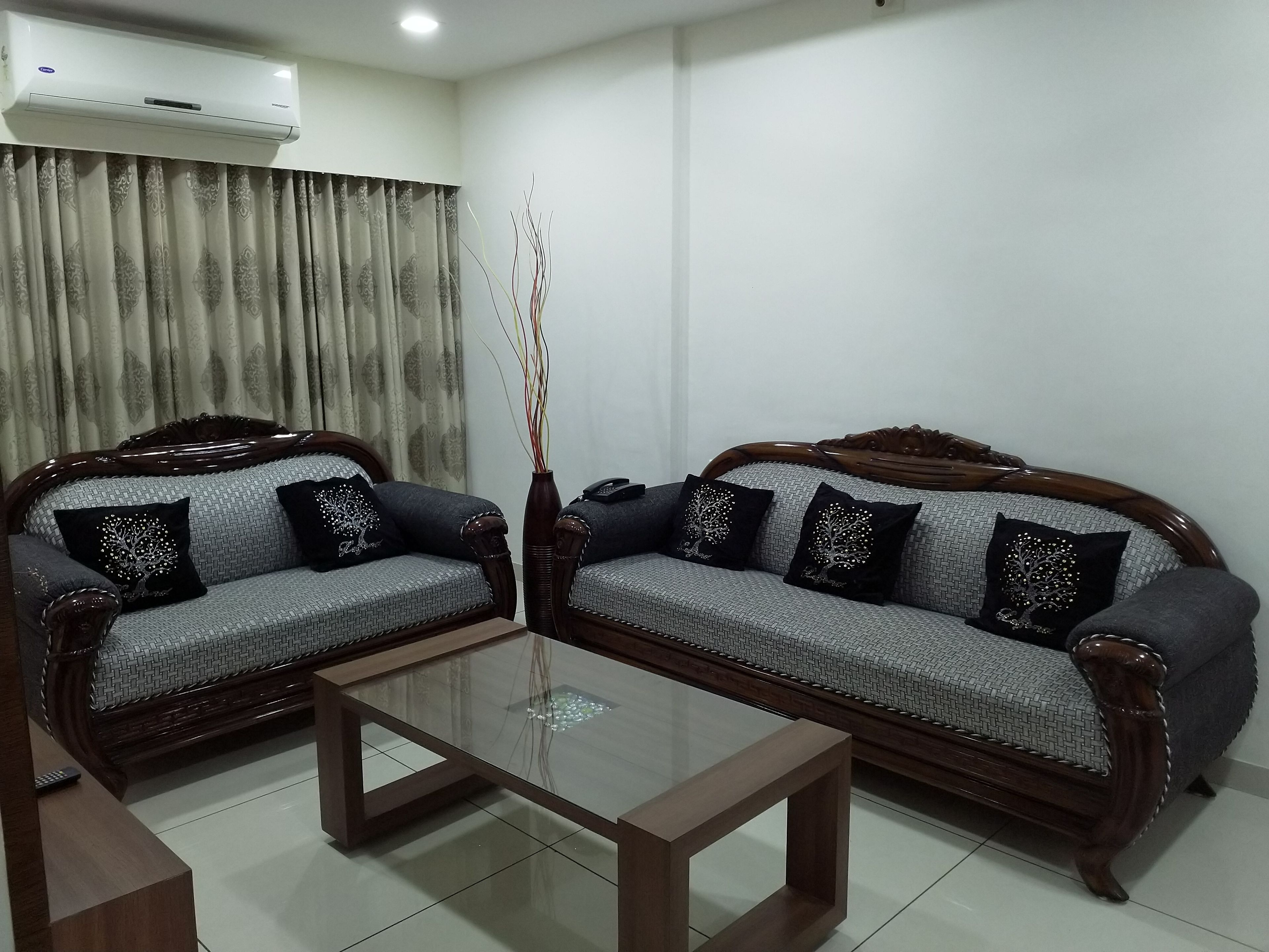 A living room with two grey sofas with wooden frames, a glass coffee table, and a wall-mounted air conditioner.