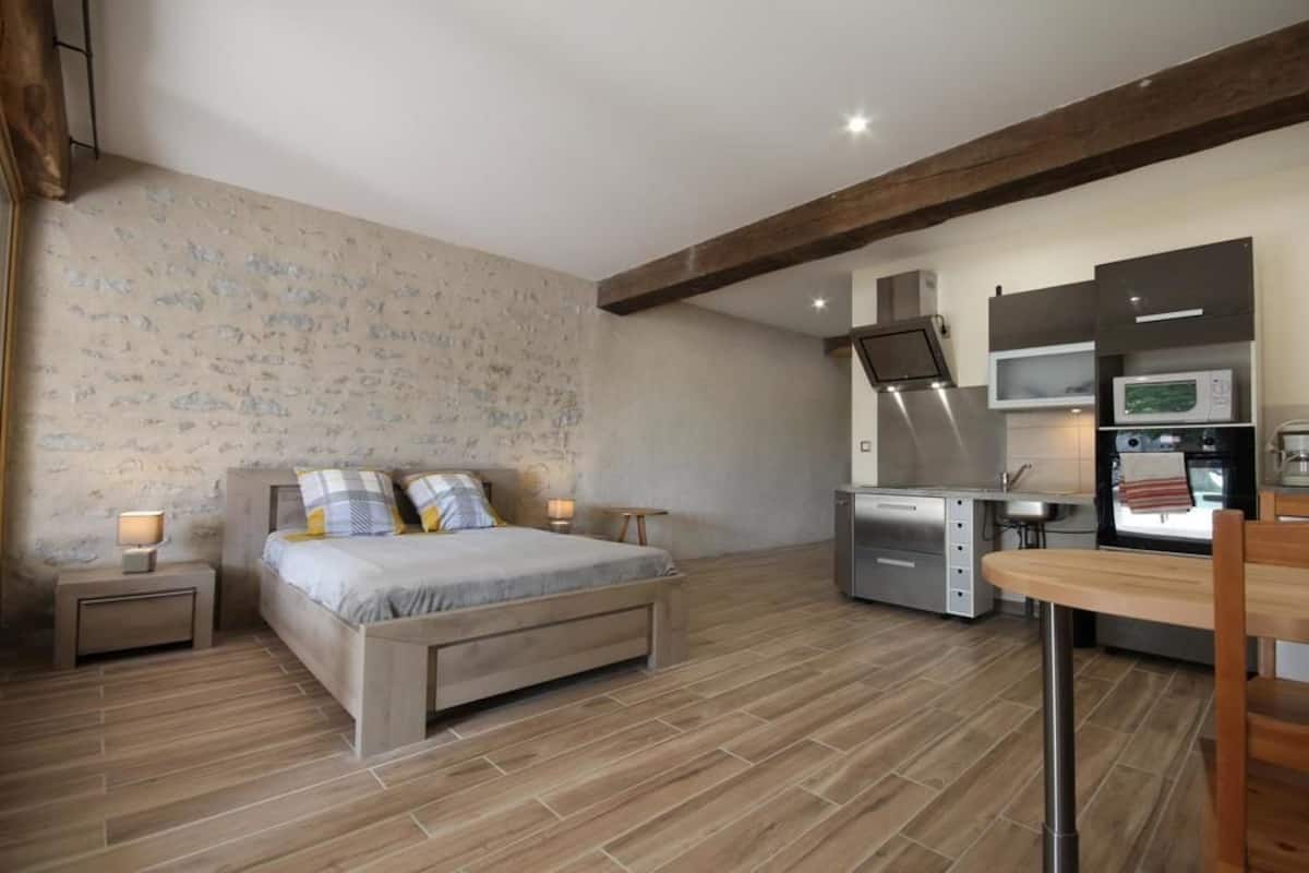 A bedroom with a bed, bedside tables, and a small desk with a chair. There is a kitchenette with a microwave, stove, and cabinets.