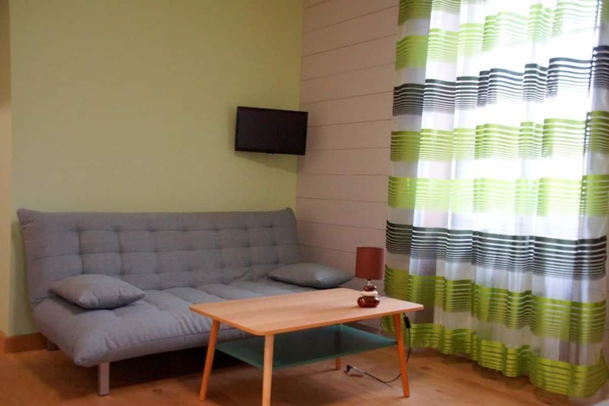 A living room with a grey sofa, a small wooden table, a TV mounted on the wall, and a curtain with green and white stripes.