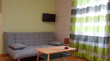 Un salón con un sofá gris, una pequeña mesa de madera, un televisor montado en la pared y una cortina a rayas verdes y blancas.