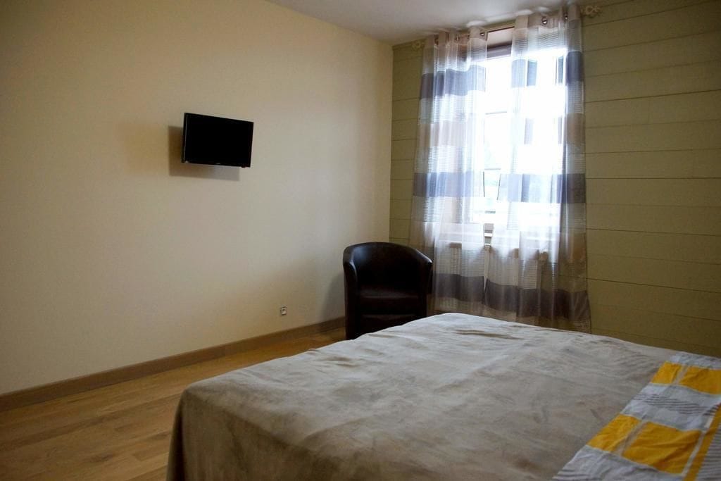 Une chambre comprenant un lit, une chaise, un téléviseur à écran plat fixé au mur et une fenêtre avec des rideaux légers.