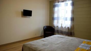 Une chambre comprenant un lit, une chaise, un téléviseur à écran plat fixé au mur et une fenêtre avec des rideaux légers.