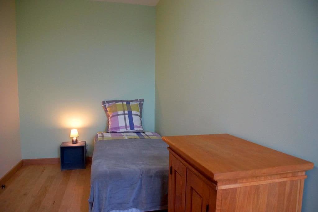 Une petite chambre avec un lit simple, une table de chevet, une lampe, un coussin à carreaux et une armoire en bois.