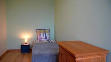 Une petite chambre avec un lit simple, une table de chevet, une lampe, un coussin à carreaux et une armoire en bois.