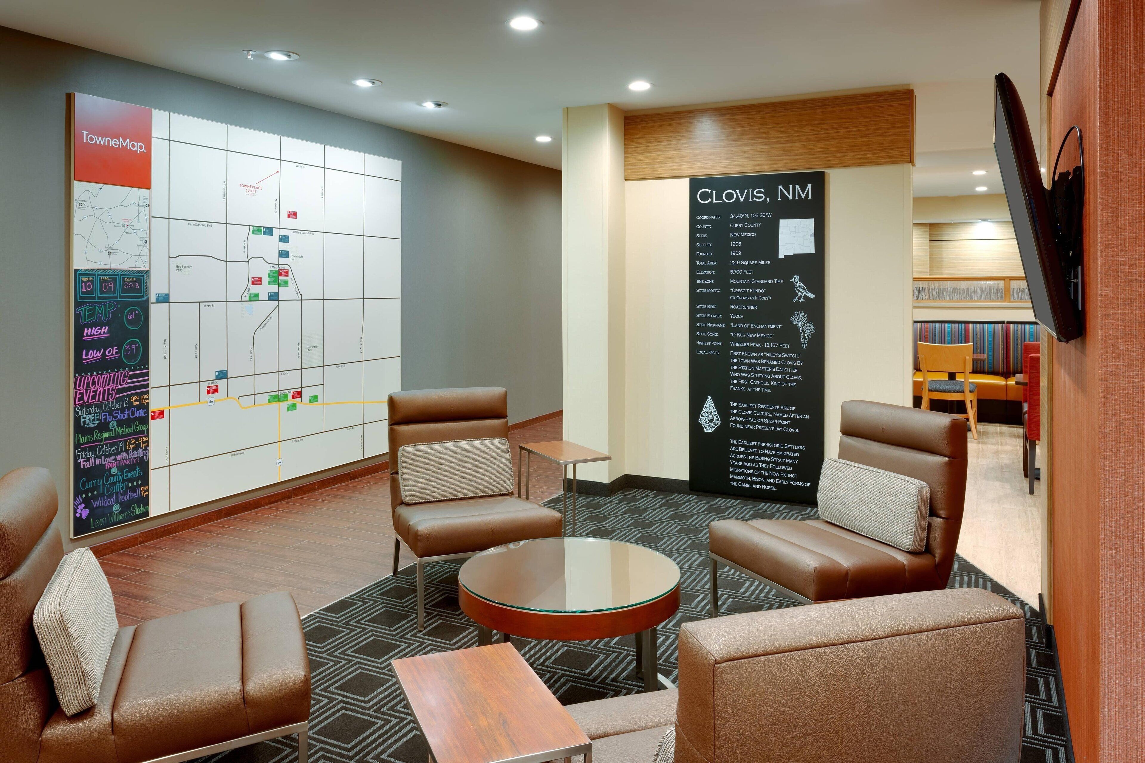 A hotel lobby with leather chairs, a round table, a map on the wall, and a sign with Clovis, NM information.