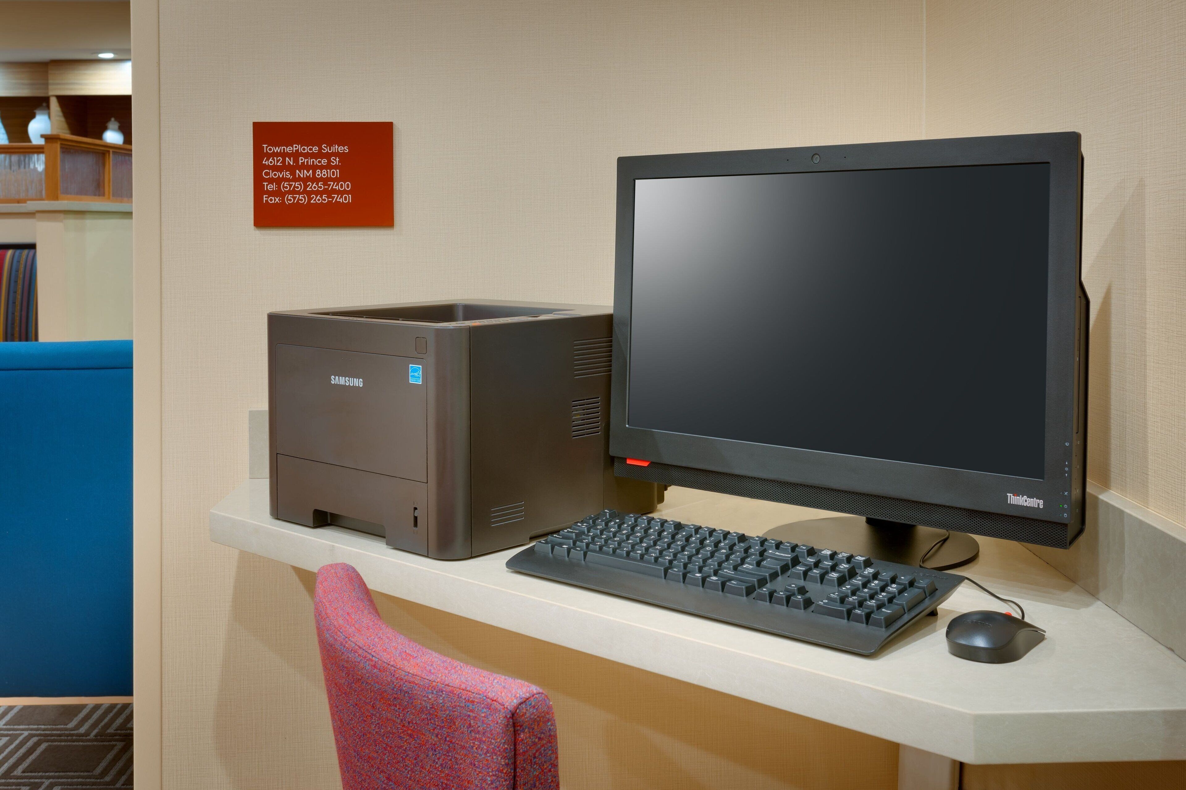 A computer setup with a monitor, keyboard, and mouse on a desk. A sign with contact information is visible on the wall.
