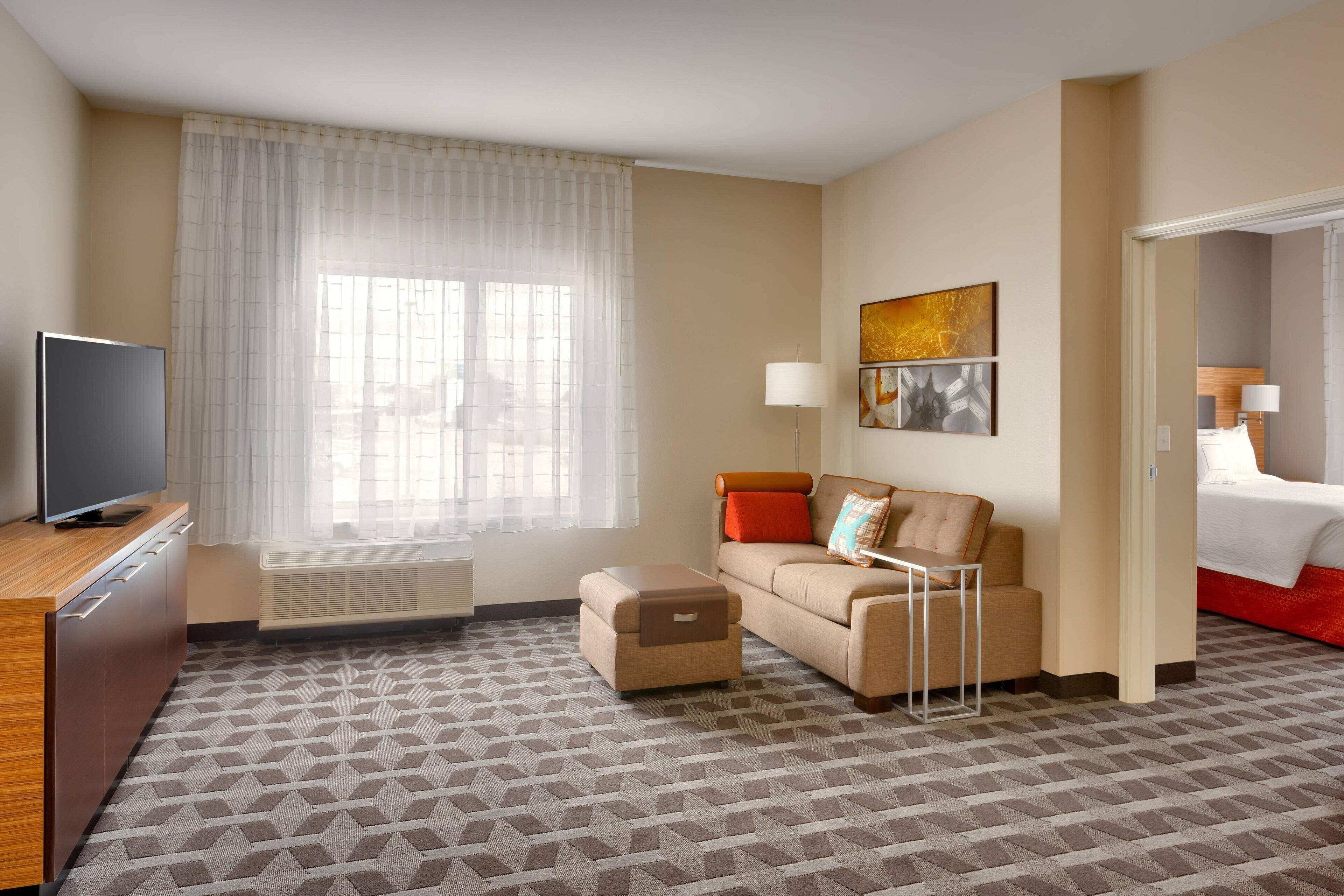 Habitación de hotel con sofá, televisión, pequeño otoman, ventana con cortinas traslúcidas y cama con detalles en rojo.