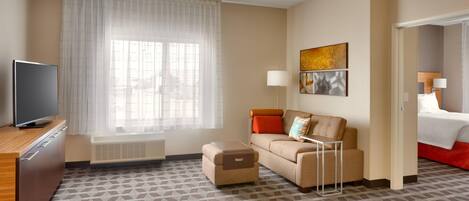 Habitación de hotel con sofá, televisión, pequeño otoman, ventana con cortinas traslúcidas y cama con detalles en rojo.