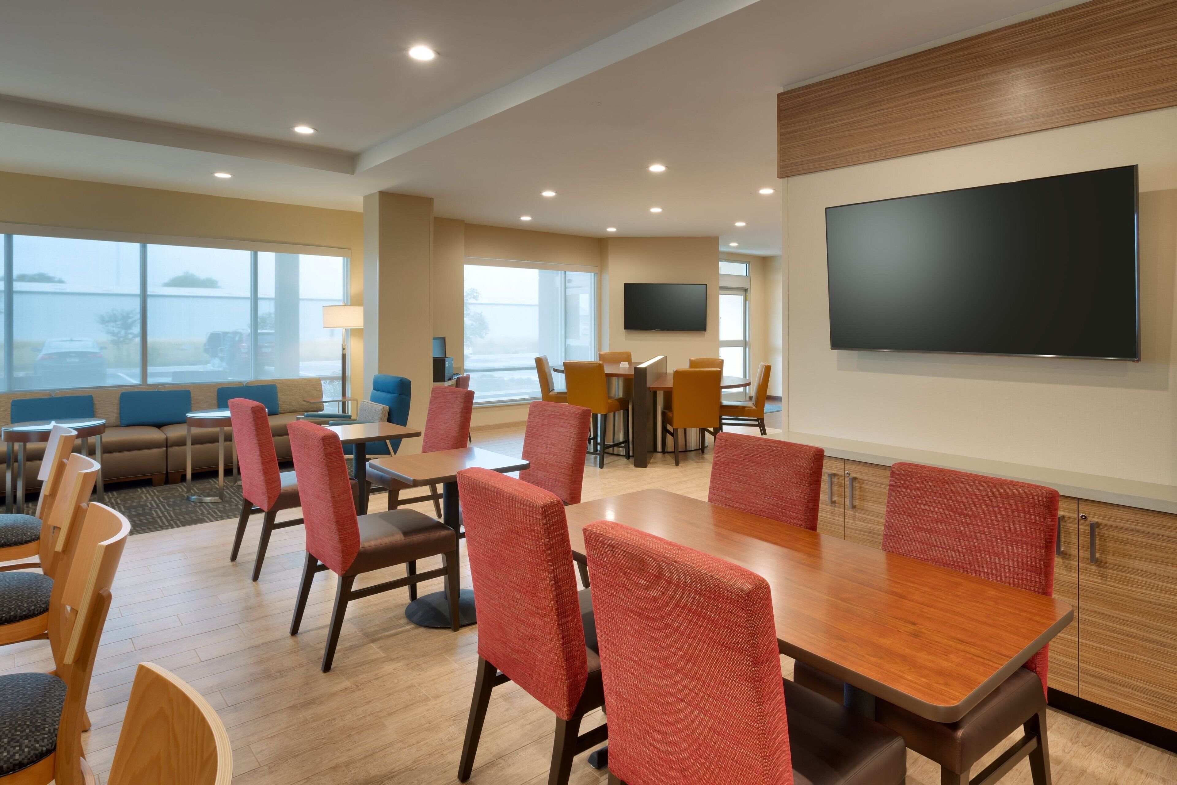 Un comedor de hotel con mesas y sillas, un televisor de pantalla plana en la pared y ventanas amplias.