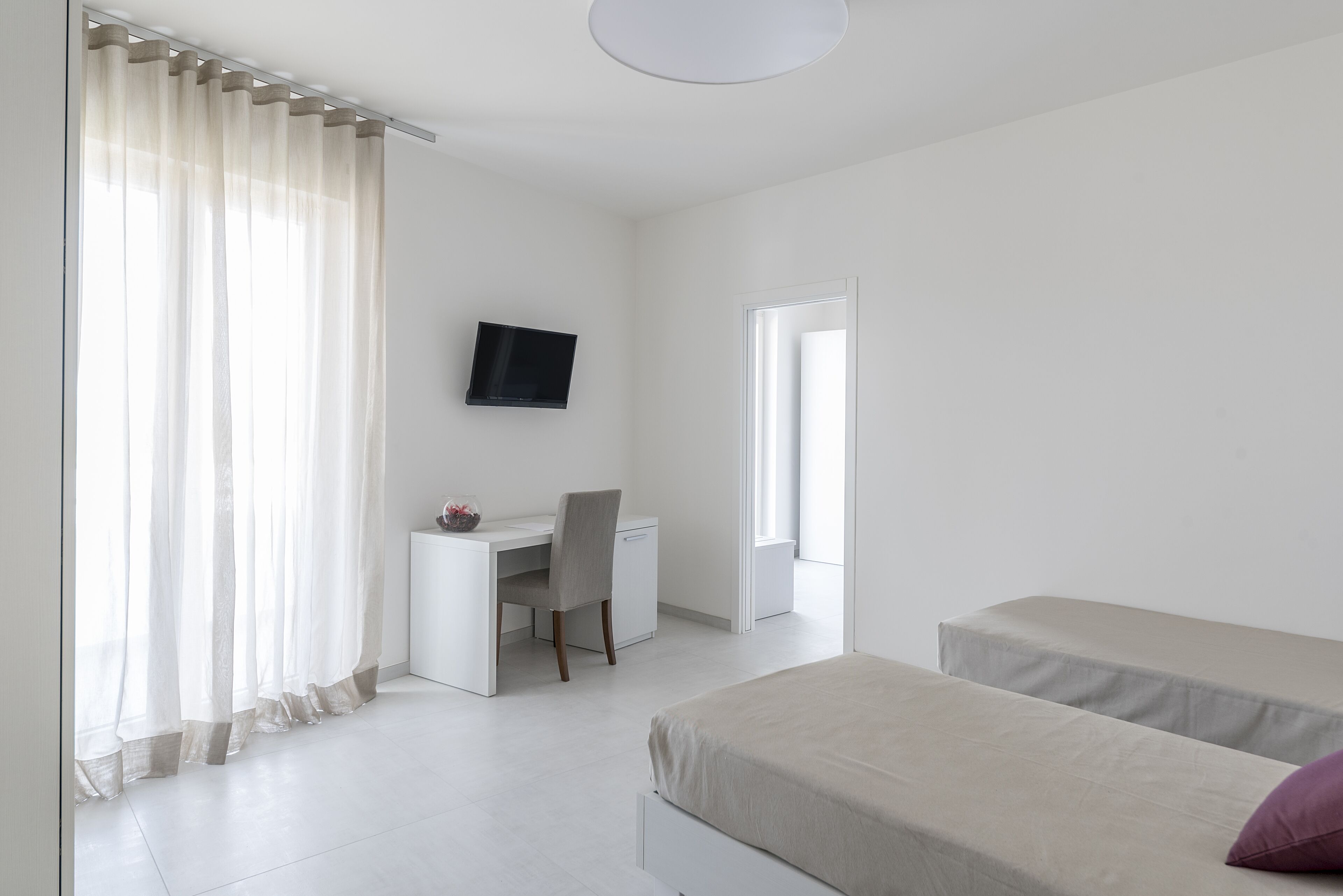 A bedroom with a desk, a bed, a TV, and a window with curtains.