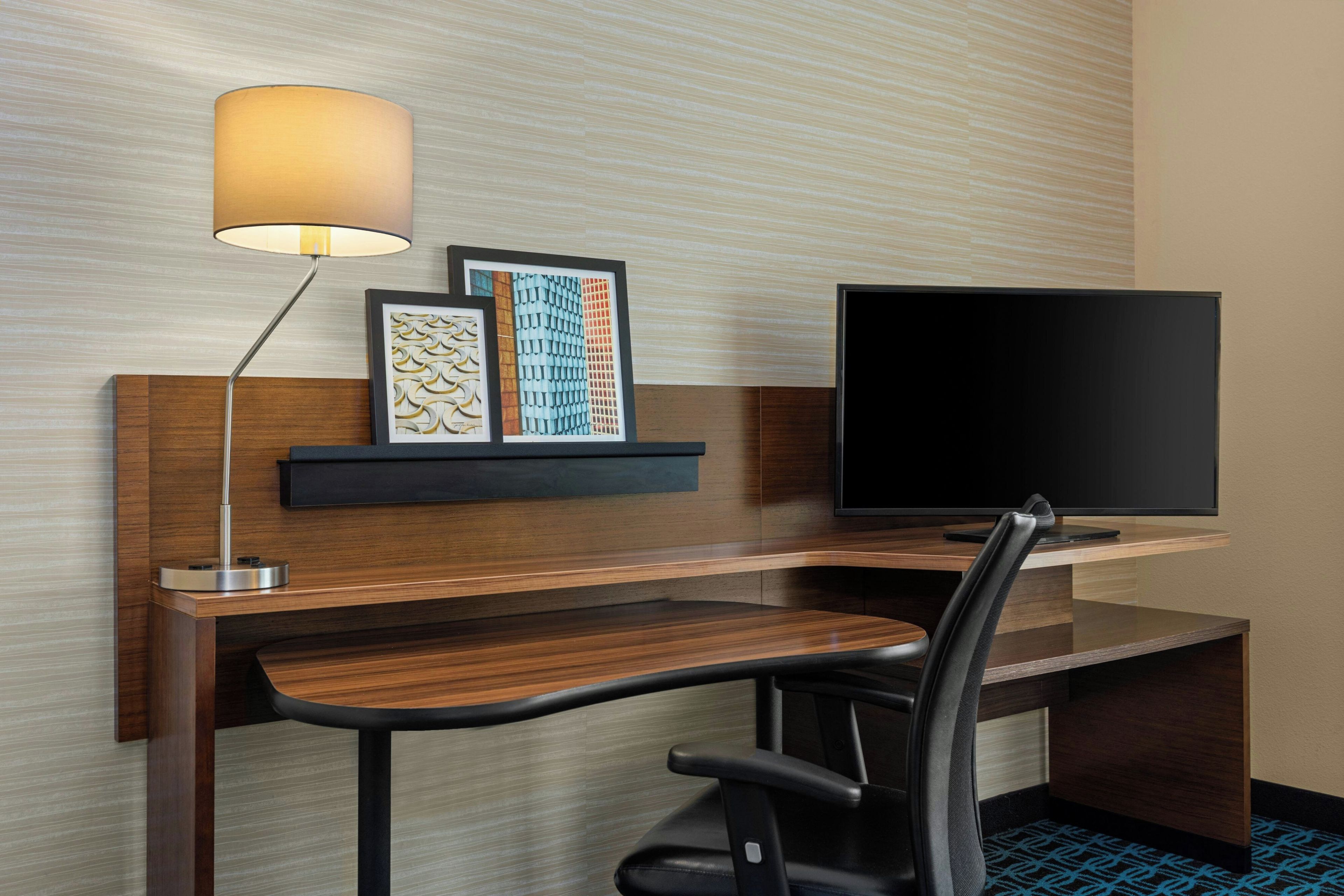 A hotel room with a wooden desk, a black office chair, a flat-screen TV, a wall-mounted lamp, and a framed artwork.