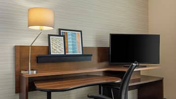 A hotel room with a wooden desk, a black office chair, a flat-screen TV, a wall-mounted lamp, and a framed artwork.