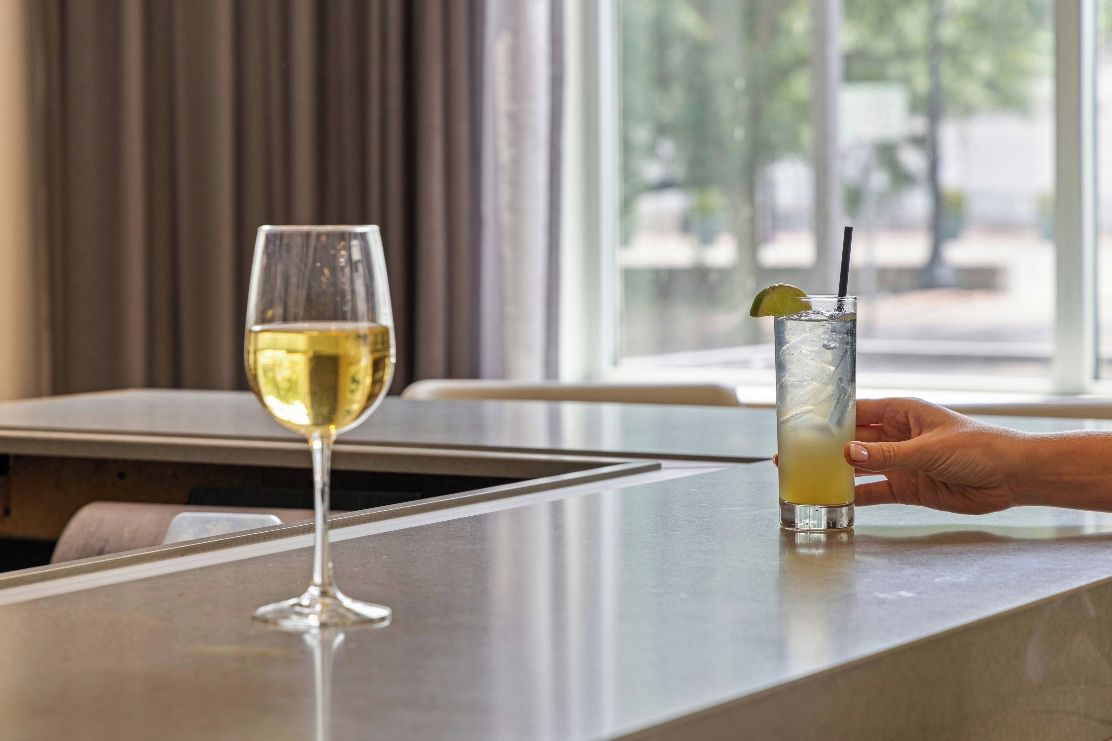 A bar area with a glass of white wine and a tall glass of a yellow beverage with a lime wedge and straw.