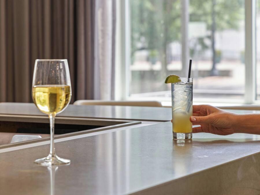 A bar area with a glass of white wine and a tall glass of a yellow beverage with a lime wedge and straw.