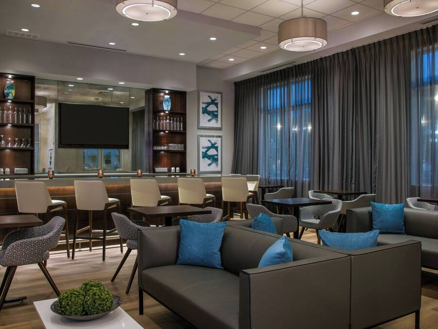 A modern hotel lobby with a bar area, seating arrangements, and a large window with curtains.