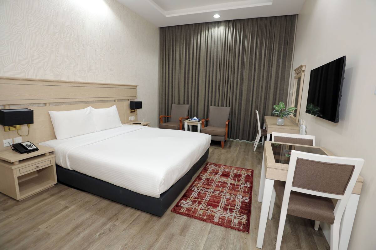 A hotel room with a bed, bedside table, lamp, chair, desk, TV, and curtains.