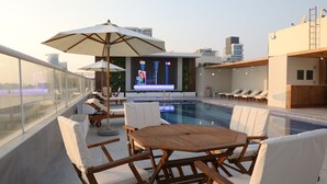 A rooftop pool area with lounge chairs, umbrellas, and a large screen displaying a show. - Wescott Plaza Hotel Apartment (Dubai)