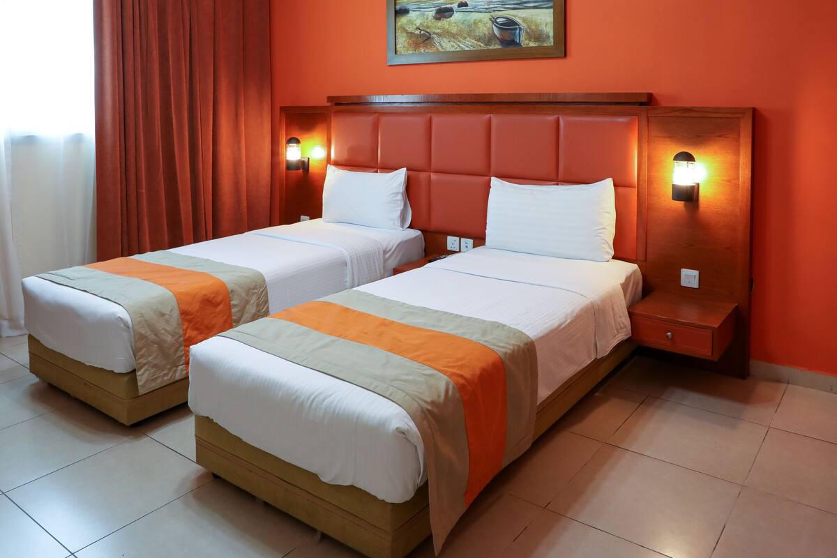 A hotel room with two single beds, a wooden headboard, a nightstand, and a painting on the wall.