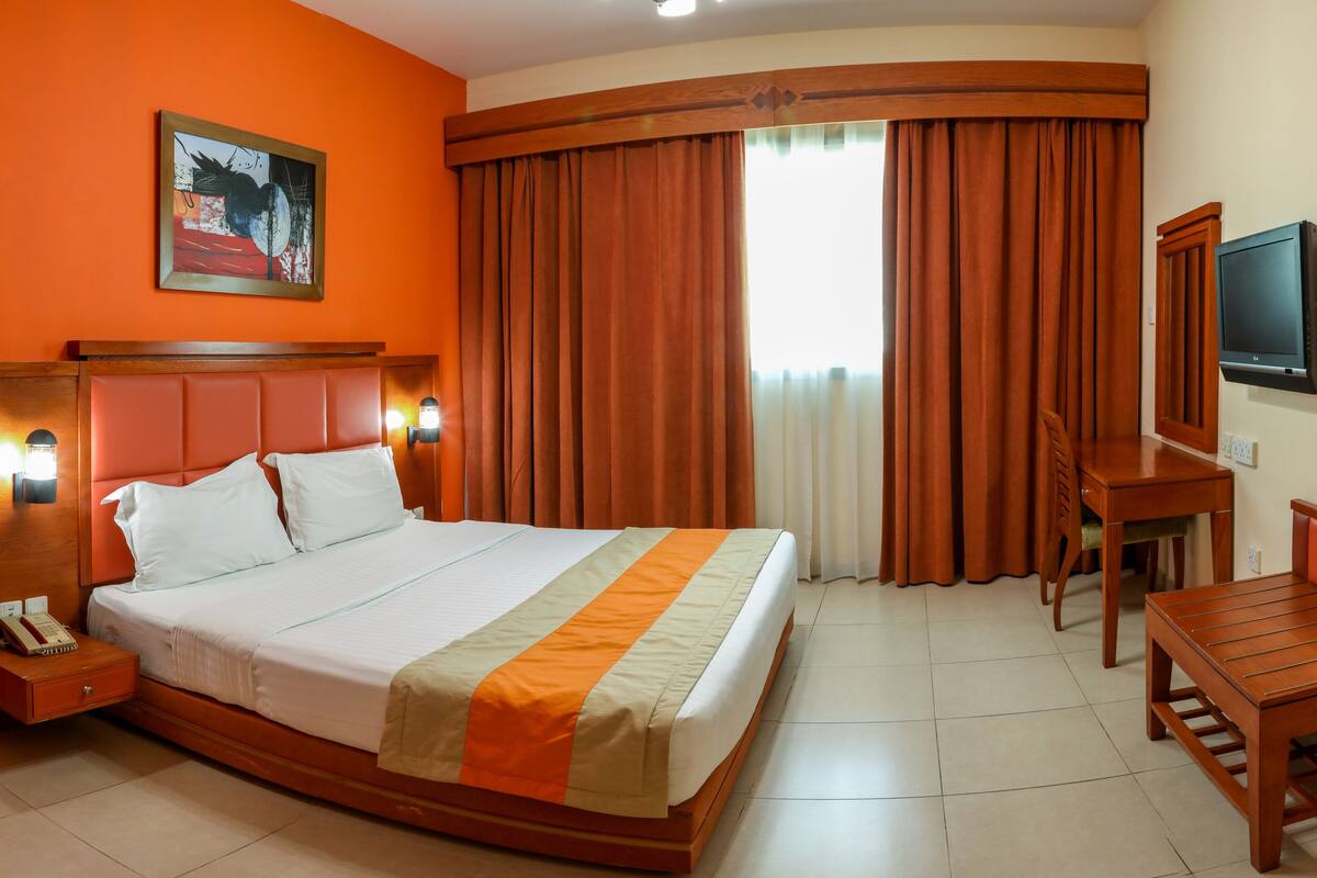 A hotel room with a bed, bedside table, lamp, TV, and a desk with a chair.