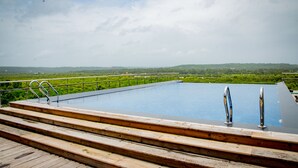 A rooftop infinity pool with wooden steps and metal railings, surrounded by lush greenery and hills. - SinQ Prive (Candolim)