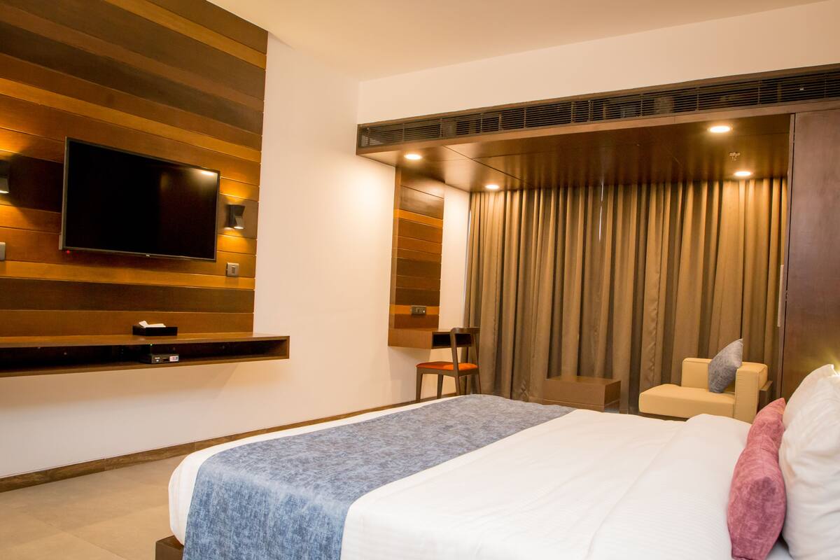 Une chambre d’hôtel moderne dotée d’un grand lit, d’un téléviseur à écran plat fixé au mur, d’un coin salon avec un fauteuil et un canapé, et d ’une grande fenêtre avec des rideaux.