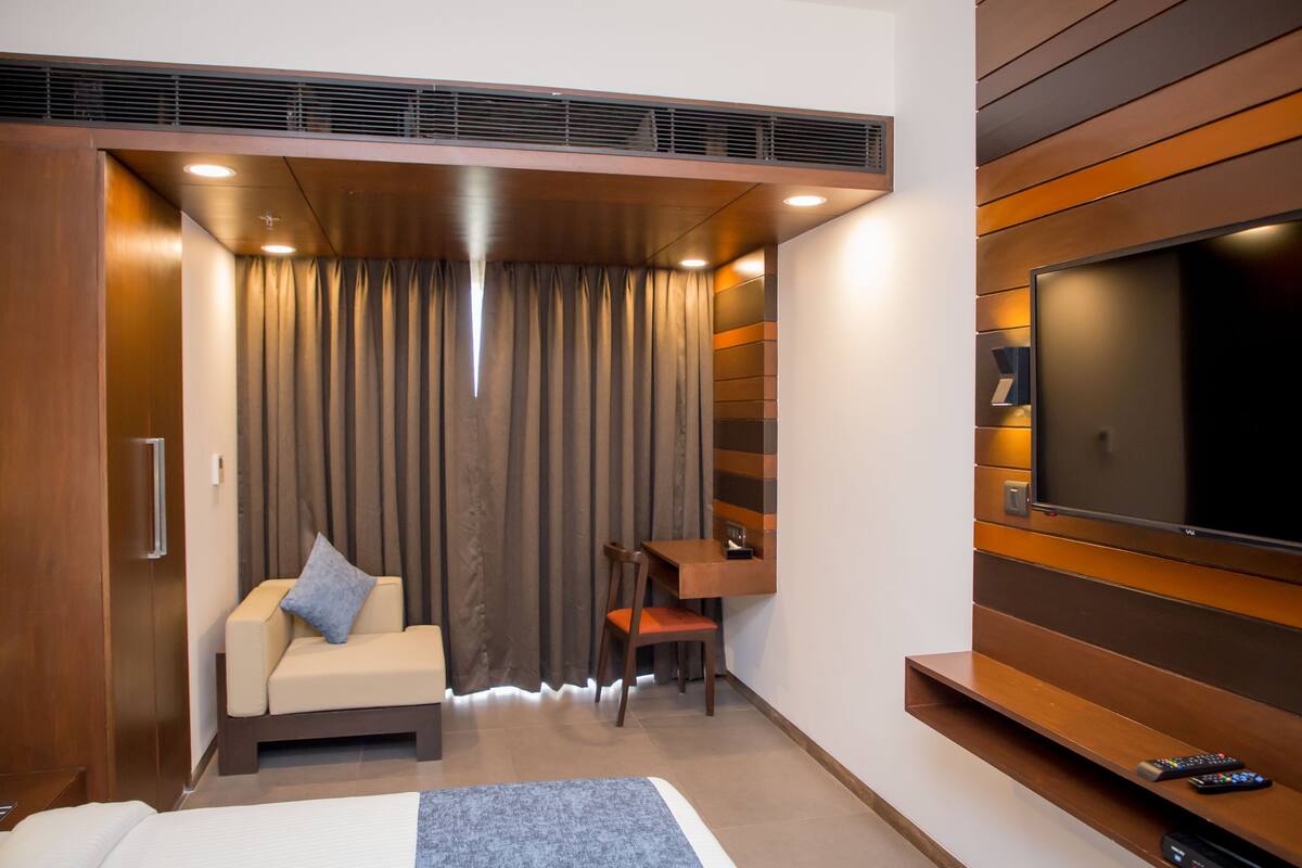 Une chambre d’hôtel comprenant un lit, un coin salon avec une chaise et une table, un téléviseur fixé au mur et une fenêtre recouverte de rideaux.