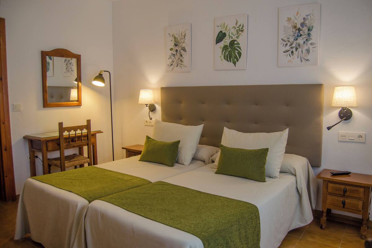 A double bed with green bedding, a wooden chair, a bedside table, and a nightstand with a lamp.
