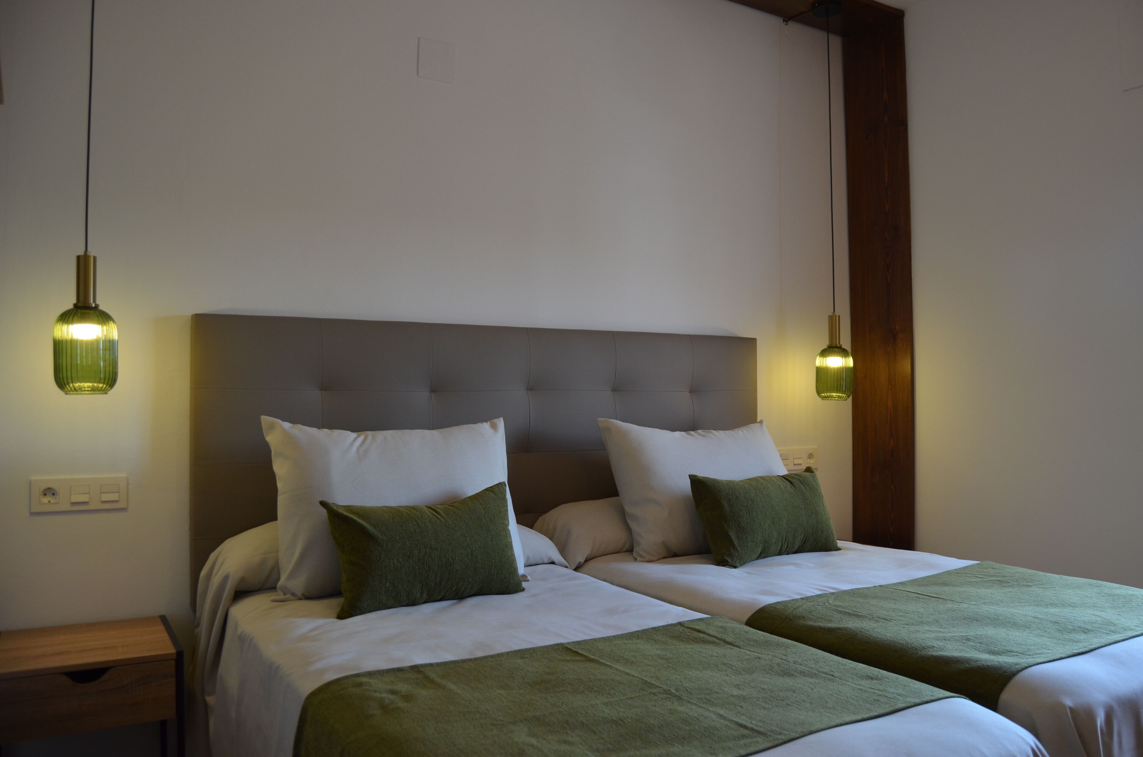 Una cama doble con cabecera gris, dos mesitas de noche y cojines decorativos verdes.