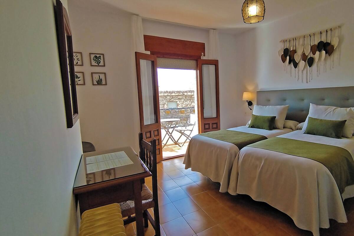 A hotel room with two beds, a desk, a chair, a balcony with a table and chairs, and a wall-mounted light fixture.