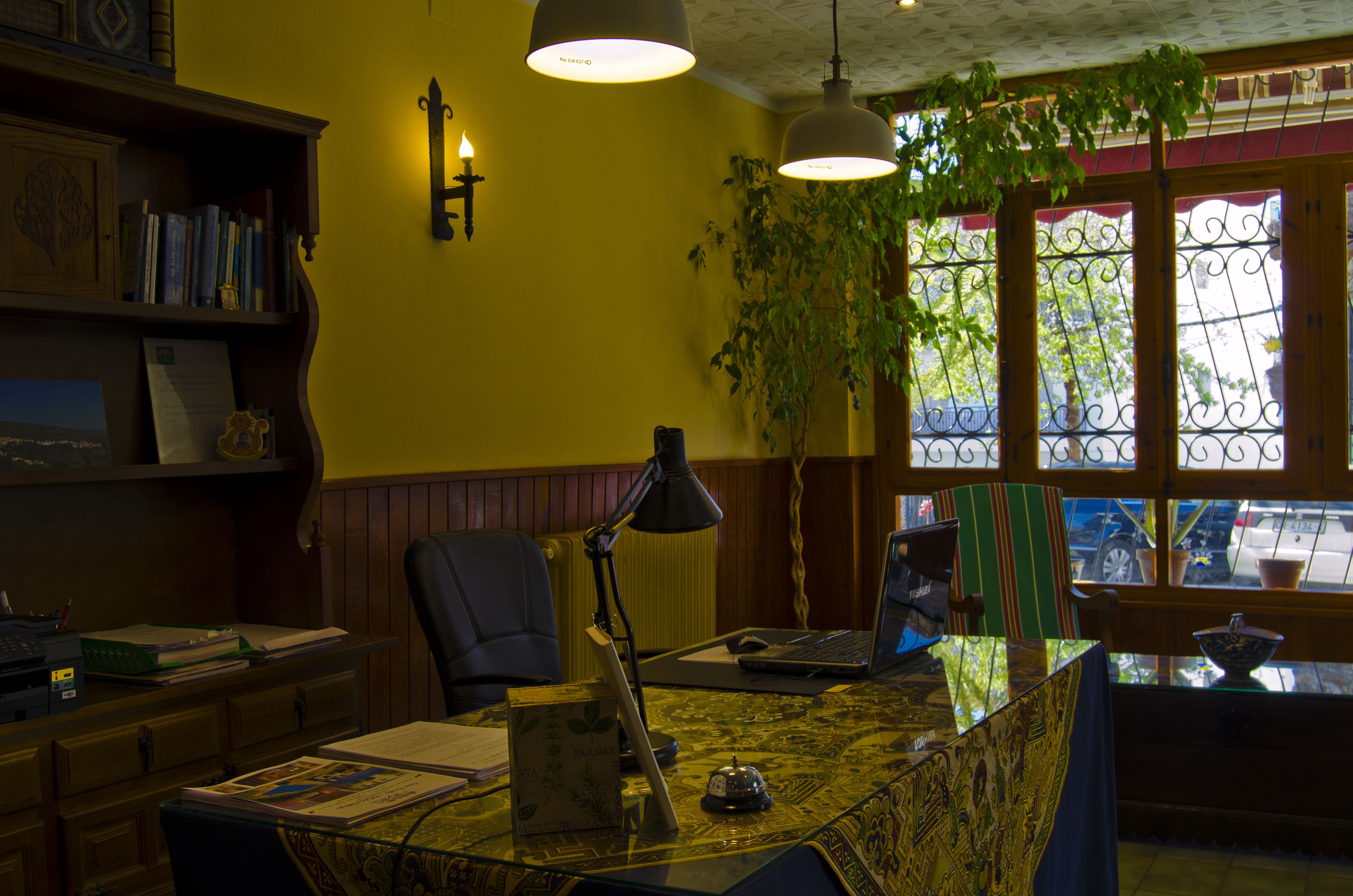A room with a desk, chair, lamp, laptop, bookshelf, and a window with a view of the street.