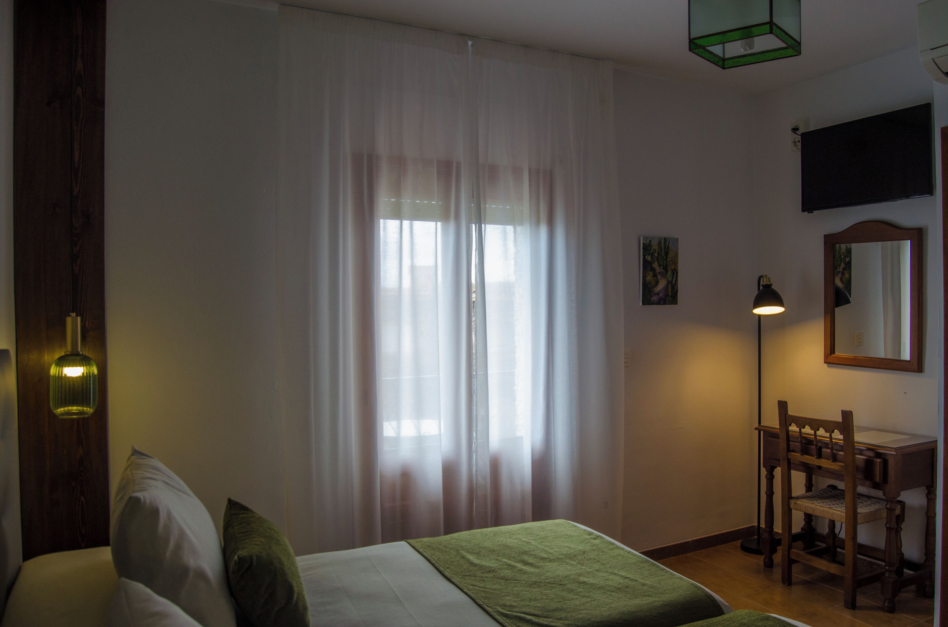 Una habitación con cama, escritorio, silla, lámpara y ventana con cortinas traslúcidas.