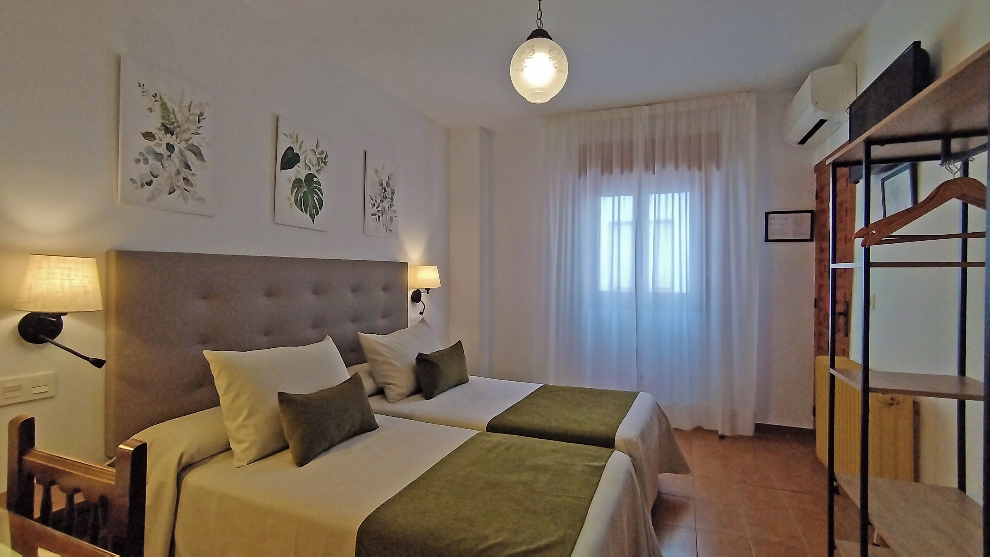 Quarto de hotel com duas camas, uma janela grande com cortinas, luminária pendente e obra de arte na parede.