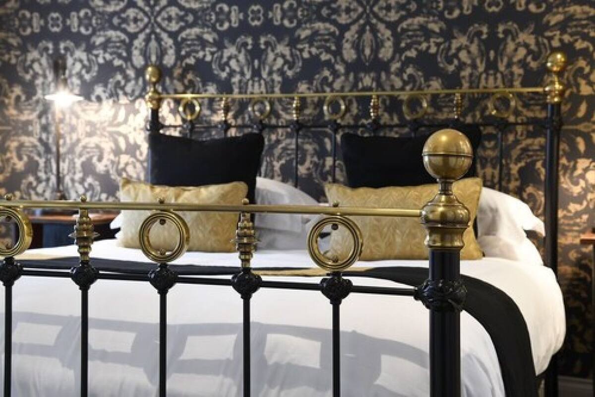 A bed with a black metal frame and gold accents, white bedding, and black and gold pillows against a patterned wallpaper background.