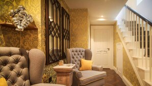 A hallway with a tufted armchair, a smaller armchair with a yellow pillow, and a decorative wall piece. A staircase with white railings is visible in the background. - Padstow Townhouse (Padstow)