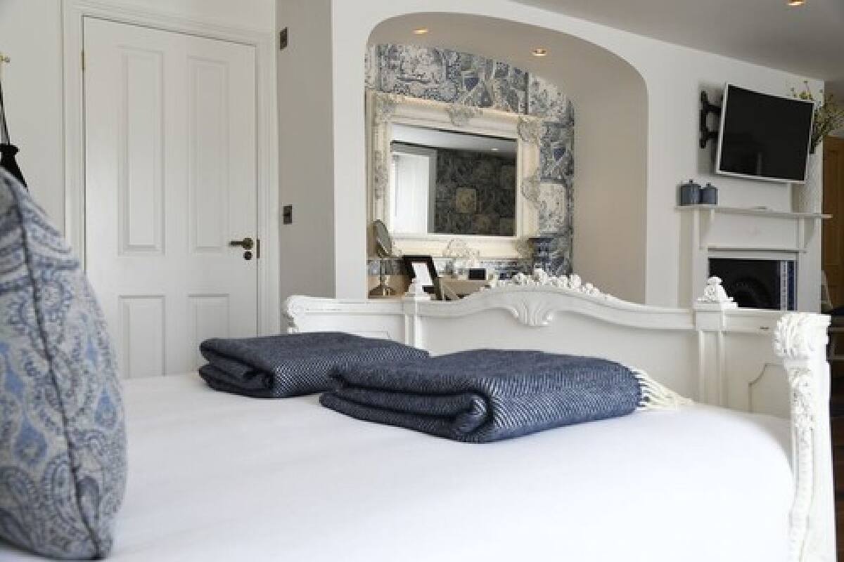 A bedroom with a white bed, two folded towels, a mirror, a television, and a fireplace.