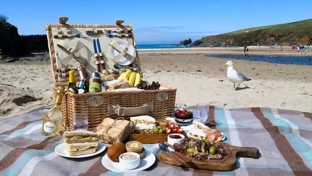 A picnic setup on a beach with a wicker basket containing food and drinks, a blanket, and a seagull nearby. - Padstow Townhouse (Padstow)