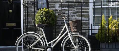 Una bicicleta blanca con una cesta, estacionada frente a un edificio de piedra con una puerta negra y un ventanal.