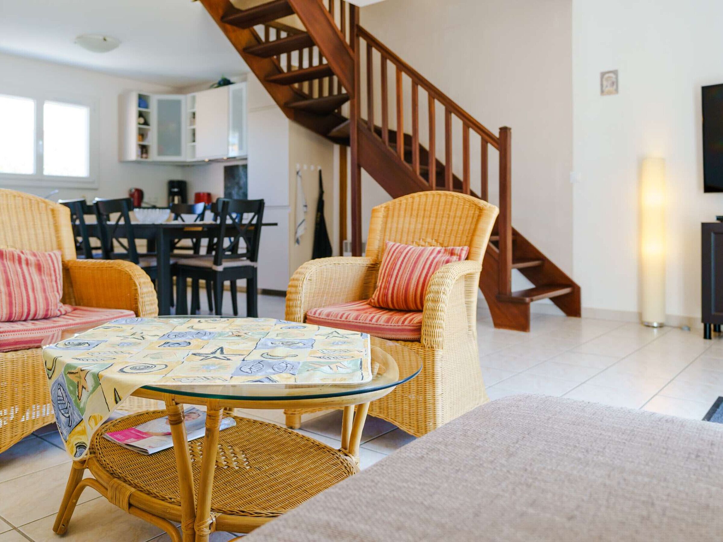 A living room with wicker furniture, a glass coffee table, and a wooden staircase.