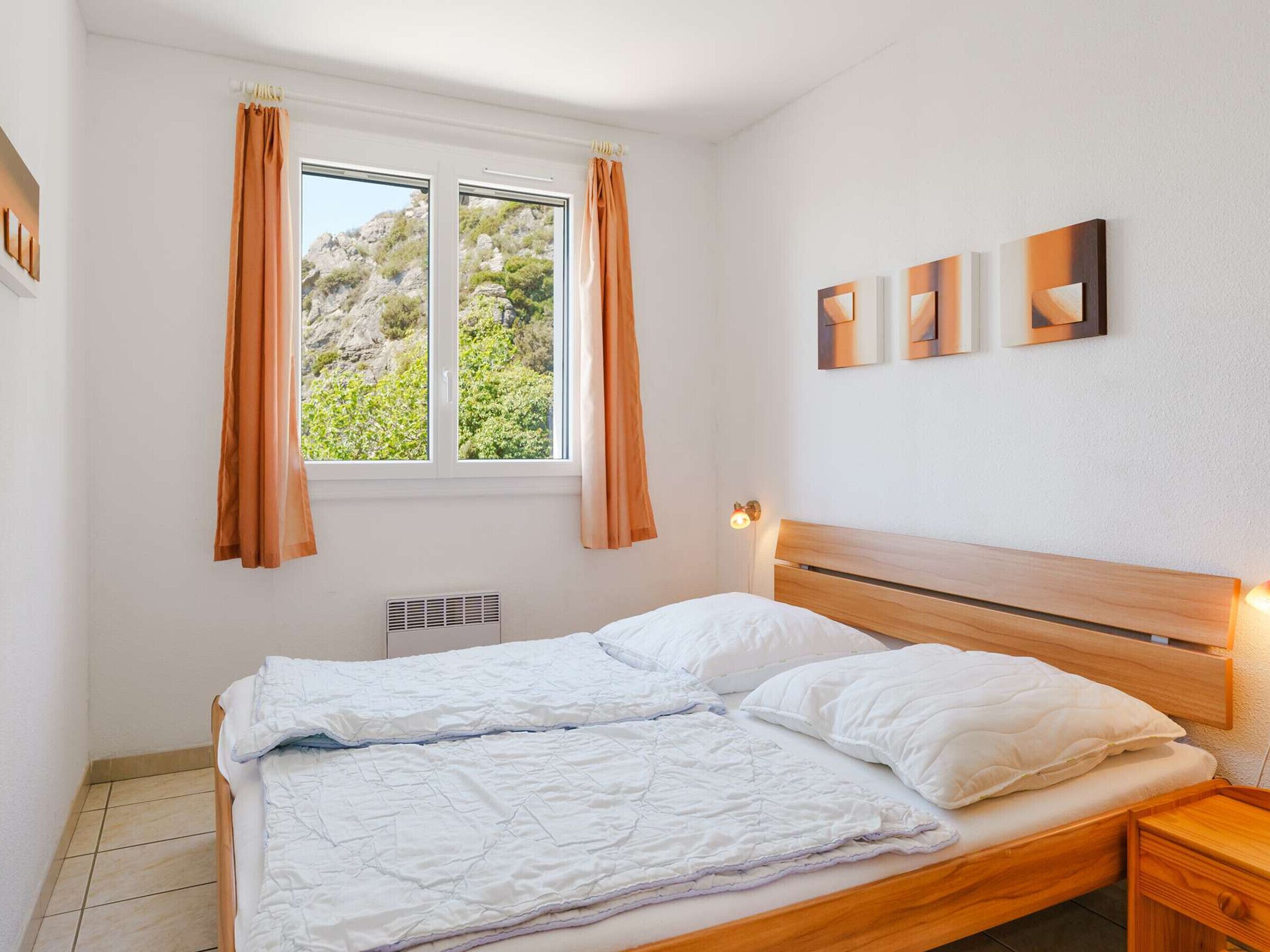 A bedroom with a wooden bed, white bedding, a window with curtains, and three wall-mounted artworks.