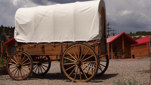 A covered wagon with wooden wheels parked on gravel, surrounded by wooden cabins with red roofs.